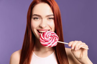 Woman with red dyed hair biting lollipop on purple background