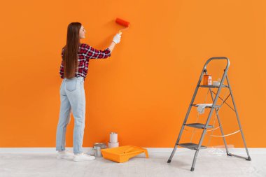 Designer painting orange wall with roller near folding ladder indoors