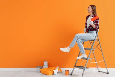 Designer with roller sitting on folding ladder near freshly painted orange wall indoors, space for text