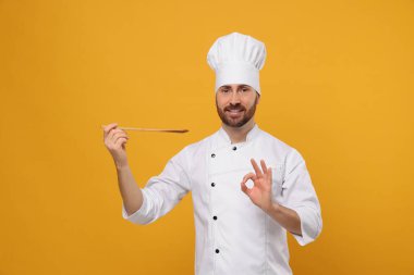 Smiling mature male chef with spoon showing ok gesture on orange background