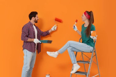 Man painting orange wall and happy woman holding can of dye with brush. Interior design