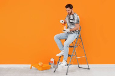 Designer with painting equipment sitting on folding ladder near freshly painted orange wall indoors, space for text