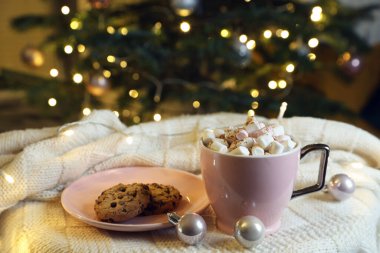 Cup of tasty cocoa with marshmallows and cookies on knitted plaid near Christmas tree indoors