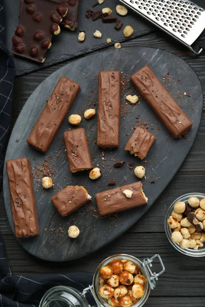 Board with delicious chocolate bars, caramel and nuts on black wooden table, flat lay