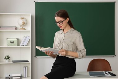 Beautiful young teacher reading book in classroom