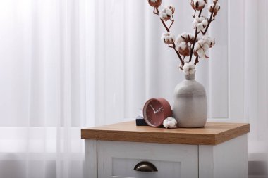 Alarm clock and cotton branches with fluffy flowers in vase on wooden commode indoors. Space for text