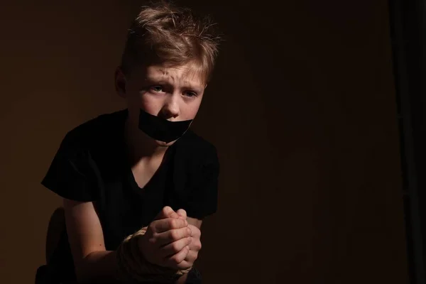 Little boy with taped mouth tied up and taken hostage against dark background. Space for text