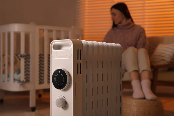 Young woman near crib and modern portable electric heater indoors, focus on device