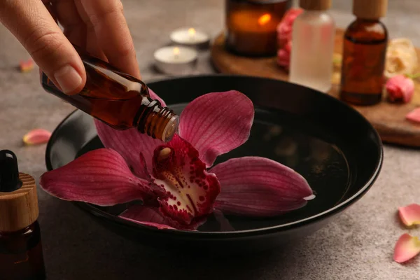 Woman dripping essential oil into bowl with flower at grey table, closeup. Aromatherapy treatment