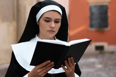 Young nun reading Bible near building outdoors