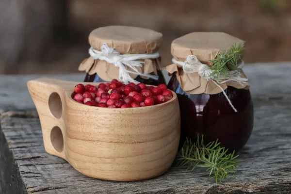 Tasty lingonberry jam in jars and cup with red berries on wooden table outdoors