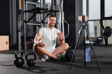 Man with ball and dumbbells streaming online training on phone at gym. Fitness coach