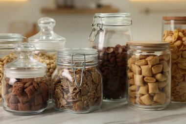 Mutfaktaki beyaz mermer masanın üzerinde farklı kahvaltı gevrekleri olan cam kaplar.