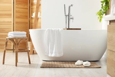 Stylish tub, wicker stool, slippers and soft towels in bathroom