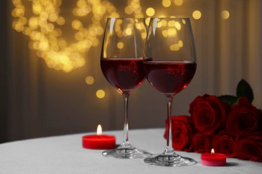Glasses of red wine, rose flowers and burning candles on white table against blurred lights, space for text. Romantic atmosphere