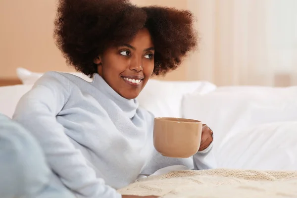 Smiling African American woman with cup of drink on bed at home. Space for text