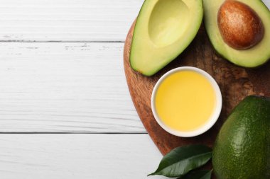 Cooking oil in bowl and fresh avocados on white wooden table, flat lay. Space for text