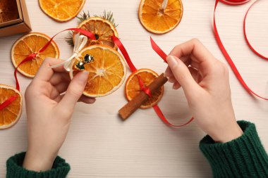 Woman making garland from dry oranges, cinnamon, ribbon and sleigh bells at white wooden table, top view