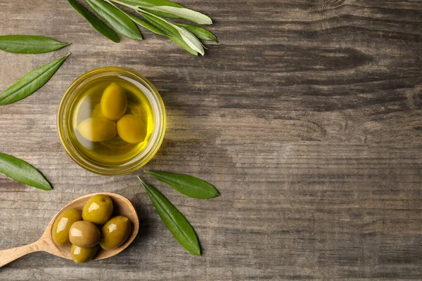 Cooking oil in bowl, olives and leaves on wooden table, flat lay. Space for text