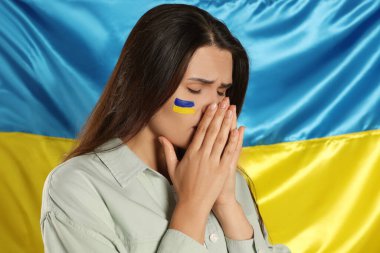 Sad young woman with clasped hands near Ukrainian flag. Space for text