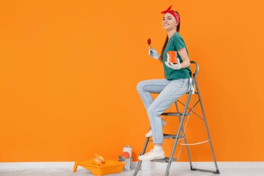 Happy designer with painting equipment sitting on folding ladder near freshly painted orange wall indoors, space for text