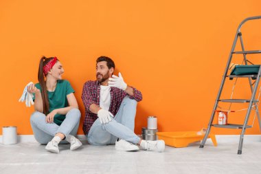 Happy designers sitting on floor with painting equipment near freshly painted orange wall indoors