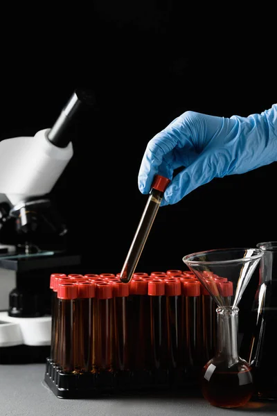 Scientist taking test tube with brown liquid from stand at grey table against black background, closeup
