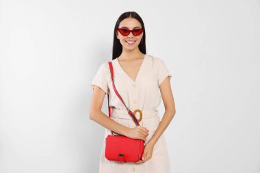 Beautiful young woman with red bag in stylish sunglasses on light grey background