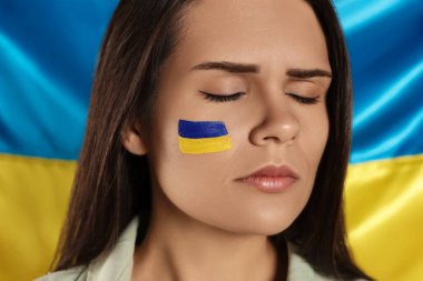 Sad young woman with face paint near Ukrainian flag, closeup
