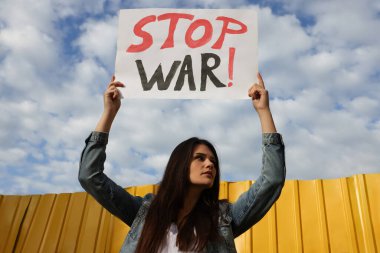 Sad woman holding poster Stop War near yellow fence under blue cloudy sky