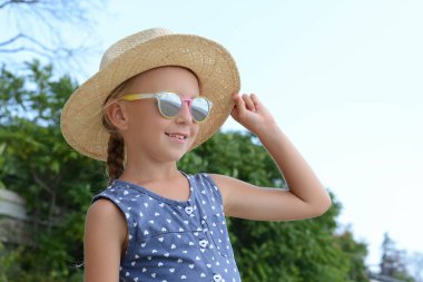 Güneşli bir günde kumsalda güneş gözlüğü ve şapka takan küçük bir kız.