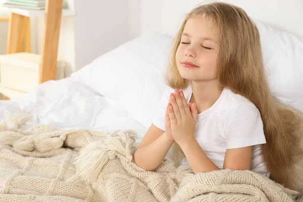 Girl with clasped hands praying on bed at home, space for text