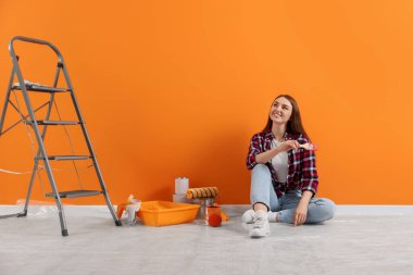 Happy designer with brush and painting equipment near freshly painted orange wall indoors