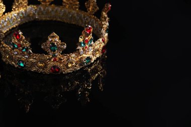 Beautiful golden crown with gems on dark mirror surface, closeup. Space for text