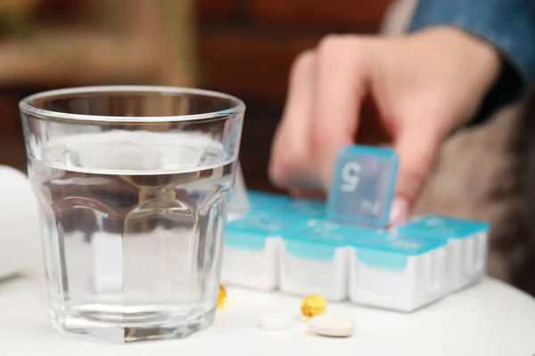 Woman taking pill from plastic box at white table indoors, focus on glass of water. Space for text