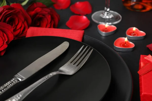 Dishware, cutlery, burning candles and bouquet on black table for romantic dinner, closeup
