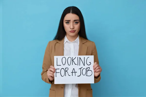 Young unemployed woman holding sign with phrase Looking For A Job on light blue background