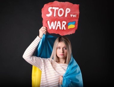 Sad woman wrapped in Ukrainian flag holding poster with words Stop the War on black background