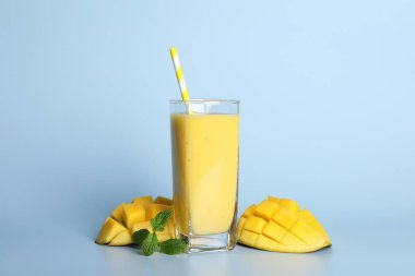 Glass of tasty smoothie with straw, mango and mint leaves on light blue background