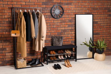 Stylish hallway with coat rack, mirror and shoe storage bench near brick wall. Interior design