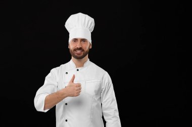 Smiling mature male chef showing thumbs up on black background, space for text