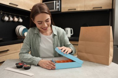 Beautiful young woman unpacking her order from sushi restaurant at table in kitchen