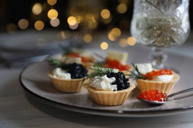 Delicious tartlets with red and black caviar served on white wooden table against blurred festive lights, closeup. Space for text