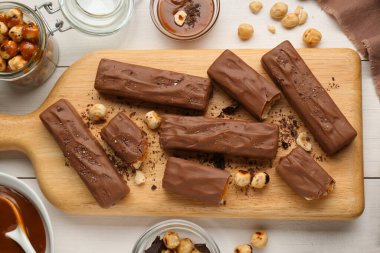 Board with delicious chocolate bars, caramel and nuts on white wooden table, flat lay