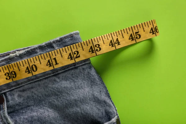 Jeans with measuring tape on green background, top view. Weight loss concept