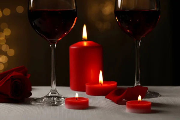 Glasses of red wine, burning candles and rose flower on grey table against blurred lights. Romantic atmosphere