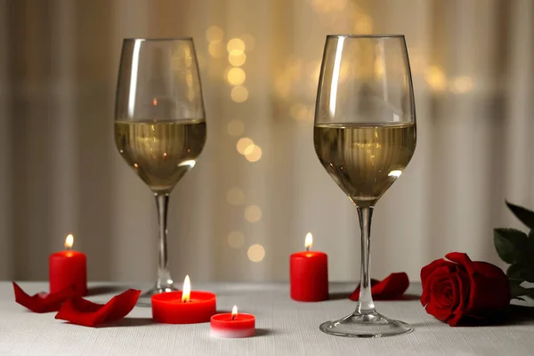 Glasses of white wine, rose flower and burning candles on grey table against blurred lights. Romantic atmosphere
