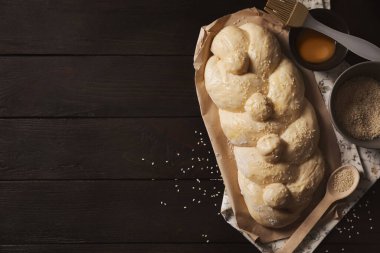 Raw braided bread, ingredients and pastry brush on black wooden table, flat lay with space for text. Traditional Shabbat challah