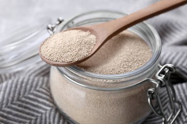 Glass jar and spoon with active dry yeast on light grey table, closeup