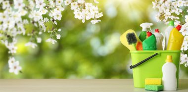Spring cleaning. Bucket with detergents and tools on wooden surface under blossoming tree against blurred green background, space for text. Banner design clipart